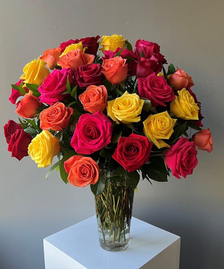 a vase filled with multi colored roses on top of a white cubed table next to a gray wall