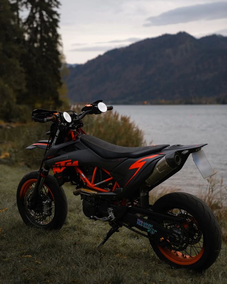 an orange and black dirt bike parked next to a body of water with mountains in the background