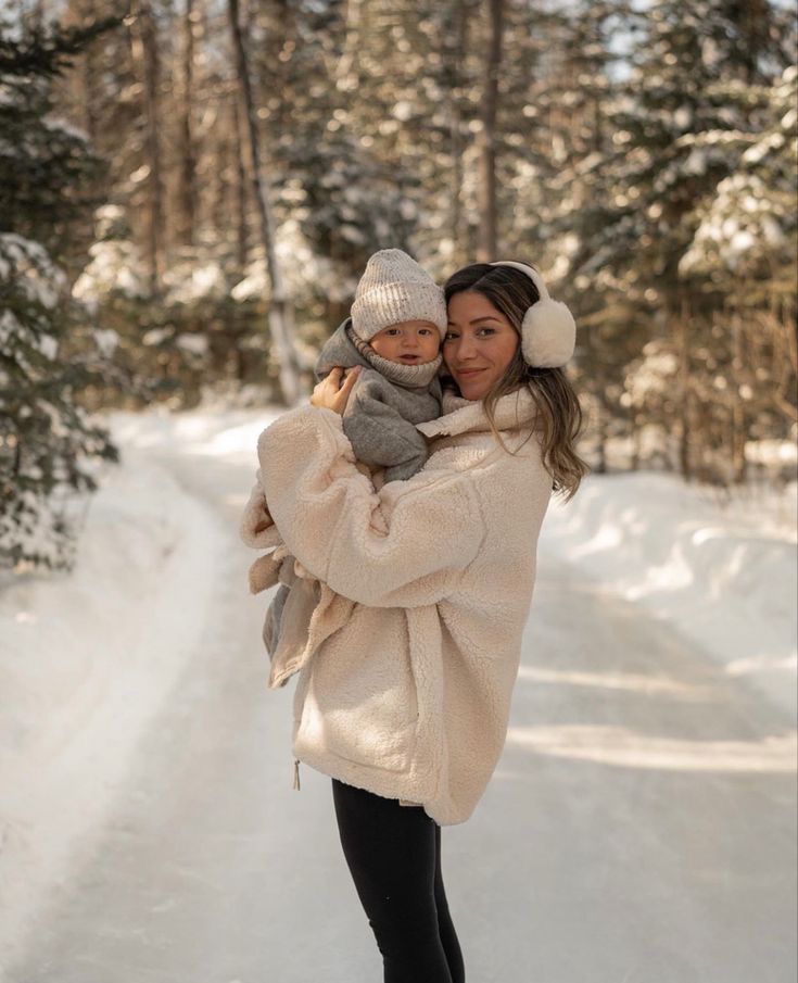 a woman holding a baby in her arms while walking down a snow covered road with trees