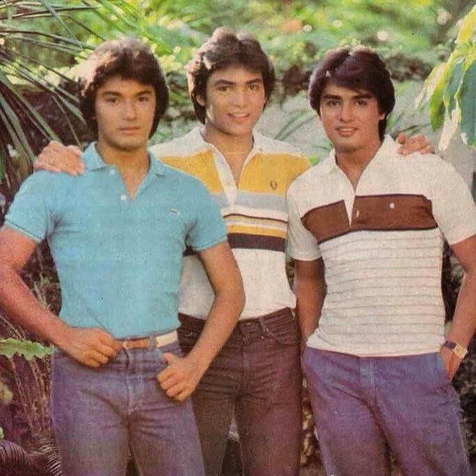 three young men standing next to each other in front of trees and bushes with their arms around one another