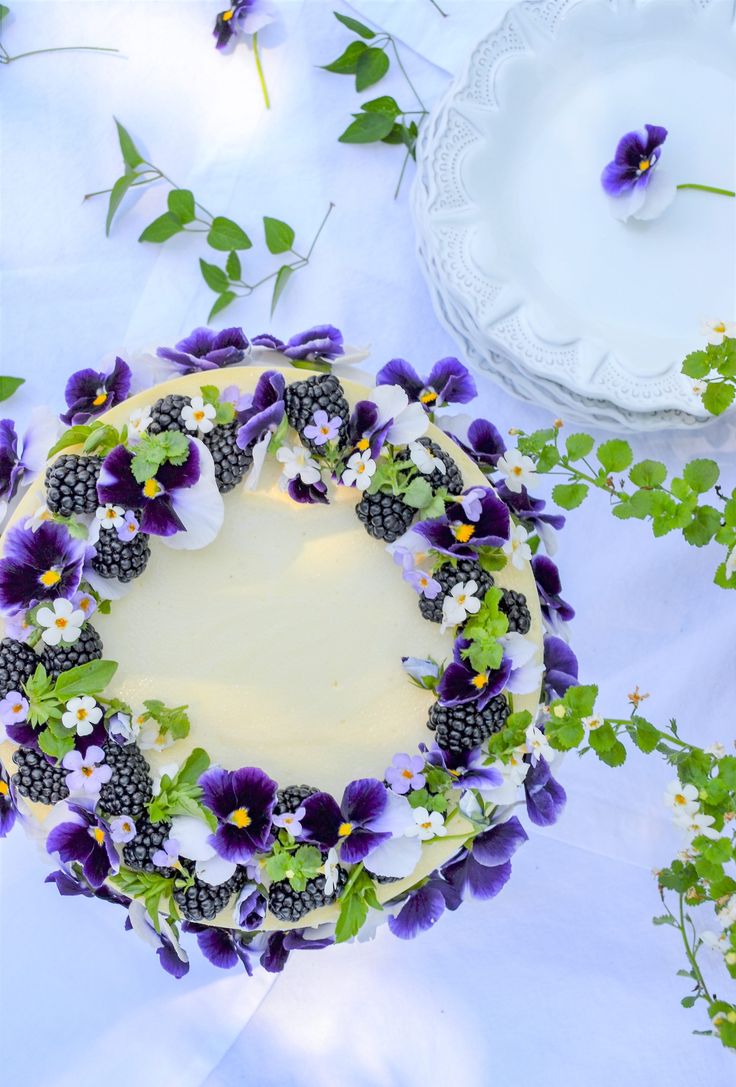 a cake decorated with purple pansies and white flowers on a blue table cloth next to a plate