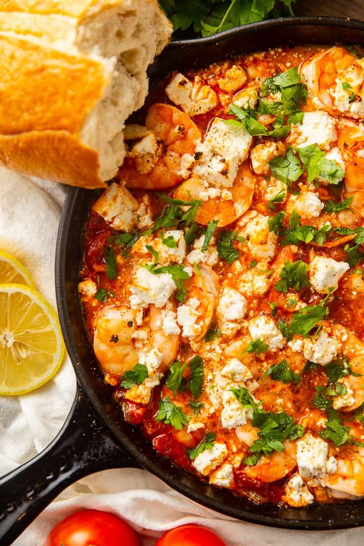 a skillet filled with shrimp and feta cheese next to sliced lemons, tomatoes and bread