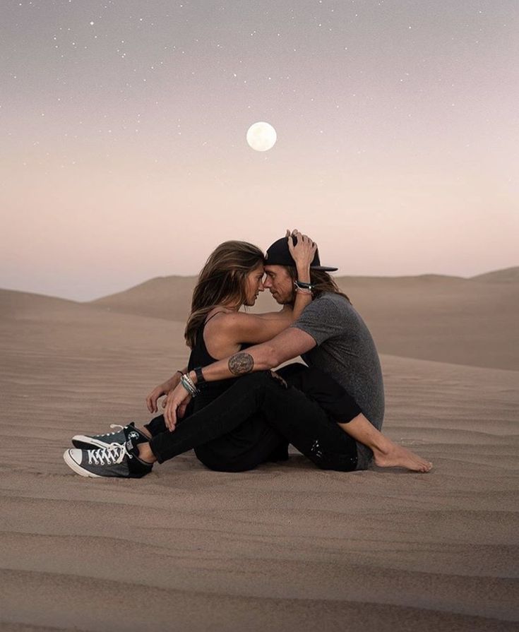 a man and woman sitting on top of a sand dune in the desert under a full moon