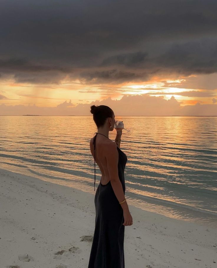 a woman standing on top of a beach next to the ocean drinking from a cup