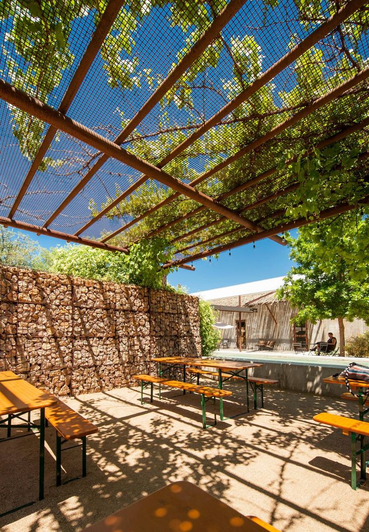 an outdoor dining area with wooden tables and benches under a pergolated roof, surrounded by greenery