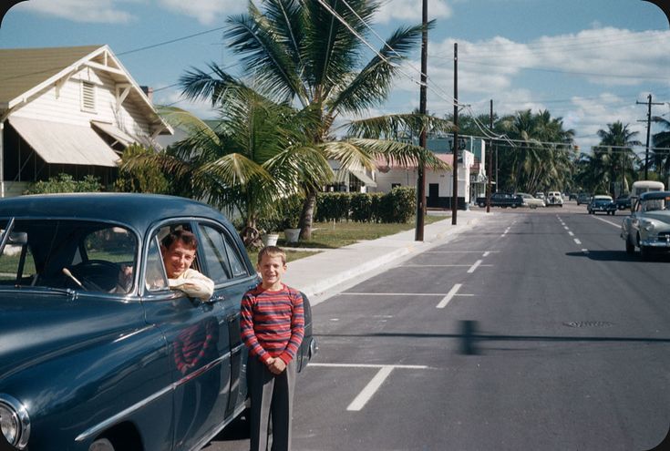 a man standing next to a blue car on a street with palm trees in the background