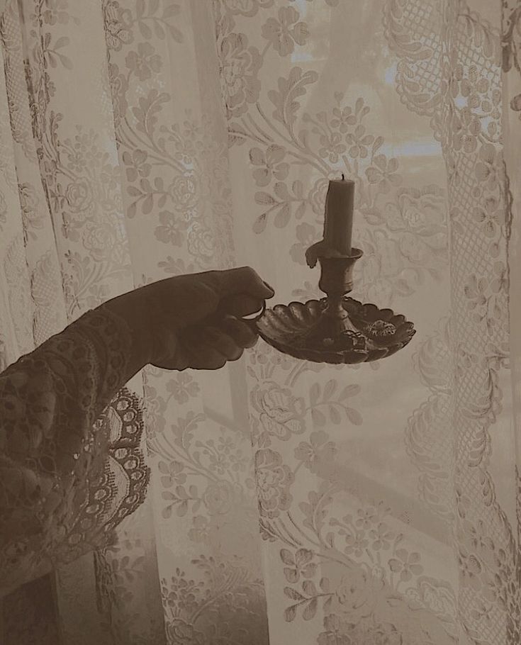 a person holding a candle in front of a window with curtains behind it and a lace curtain