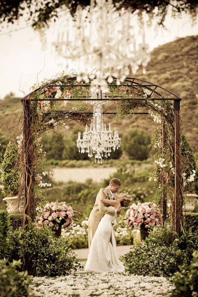 a bride and groom kissing under an outdoor wedding ceremony arch with chandelier in the background