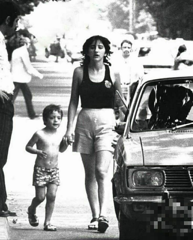 a woman and child are walking down the street with an older car in front of them