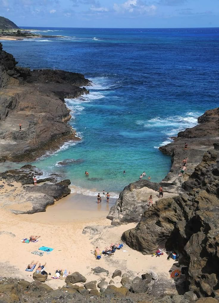 people are on the beach and in the water near some rocks with waves crashing against them