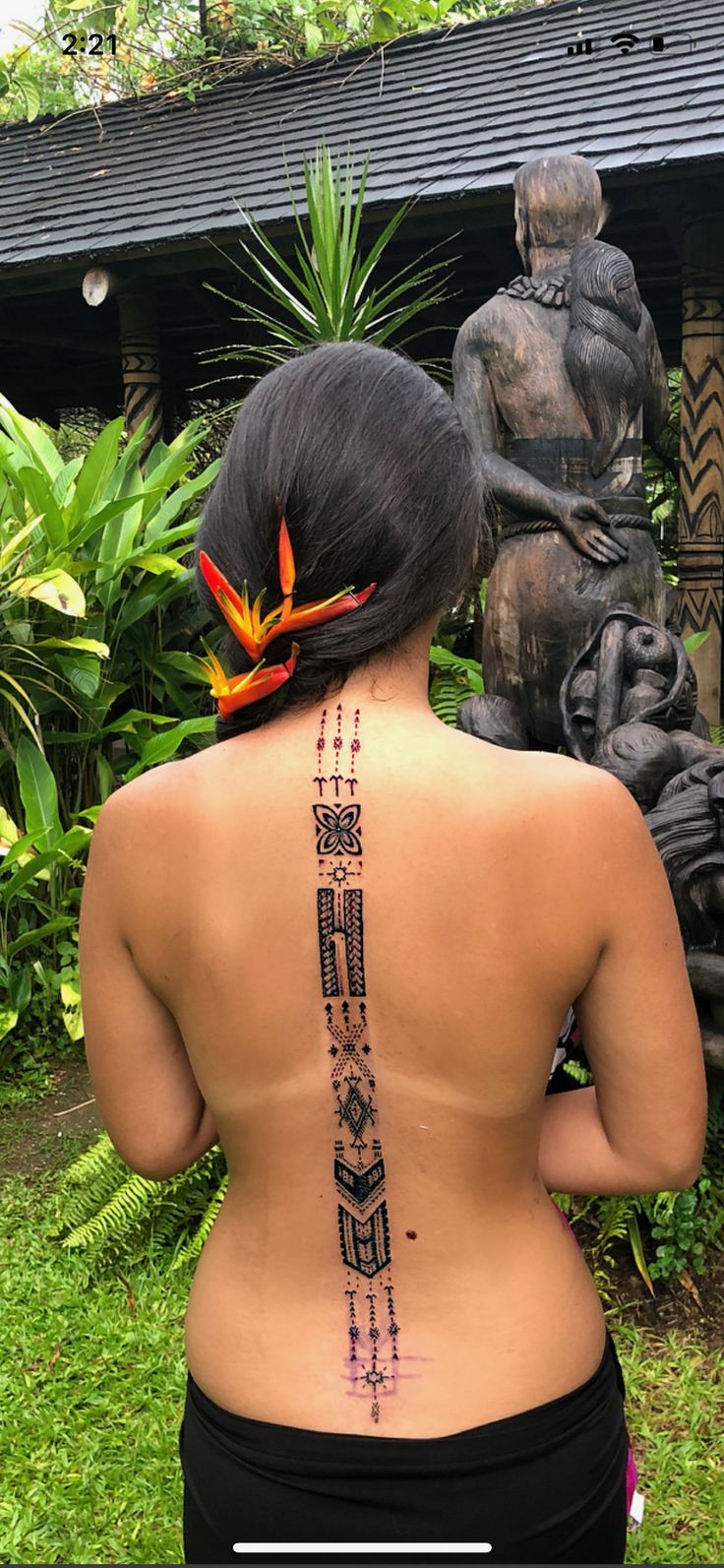 a woman with a tattoo on her back standing in front of a statue and flowers