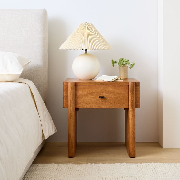 a nightstand with a lamp on top of it next to a white bed in a bedroom