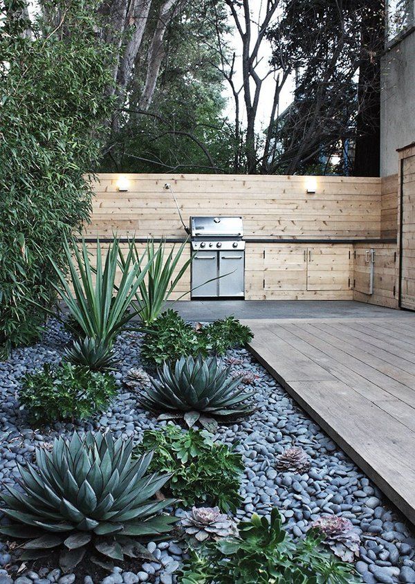 an outdoor kitchen is surrounded by plants and rocks, along with a wooden deck that leads to the back yard