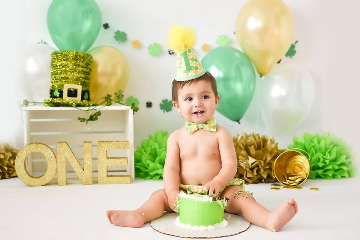 a baby sitting on the floor with a cake and balloons in the background at a st patrick's day party