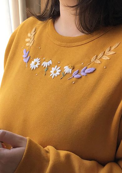 a woman wearing a yellow sweater with flowers embroidered on the chest and sleeves, standing in front of a window