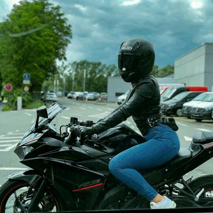 a woman riding on the back of a black motorcycle down a street next to a parking lot