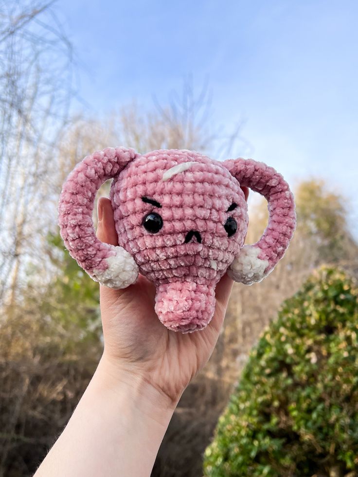 a pink crocheted elephant with white tusks and ears is held up in the air by someone's hand