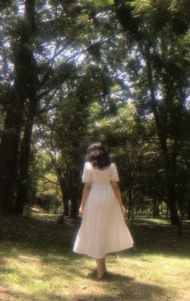 a woman in a white dress is walking through the woods with her back to the camera