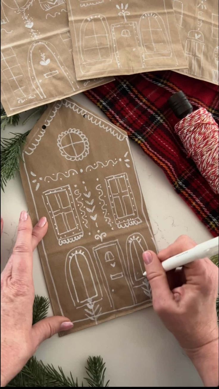 two hands are decorating a gingerbread house on a sheet of brown paper with white lettering