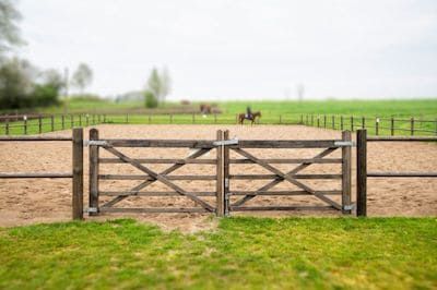 an open gate in the middle of a dirt field with horses grazing on grass behind it