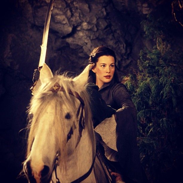 a woman riding on the back of a white horse next to a stone wall and trees