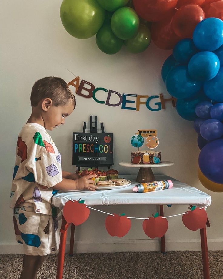 a young boy standing in front of a table with a cake on it and balloons behind him