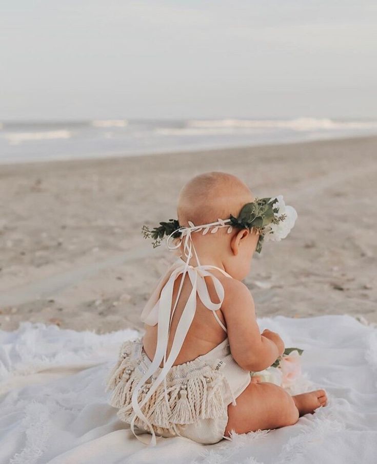 a baby sitting on the beach wearing a white dress with flowers in it's hair