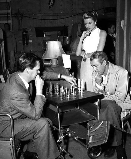 two men playing chess in a room full of other people and one man sitting at a table