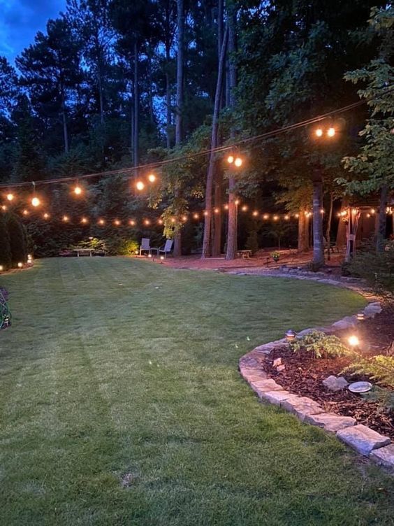 a backyard with lights strung over the grass and trees in the background at night time