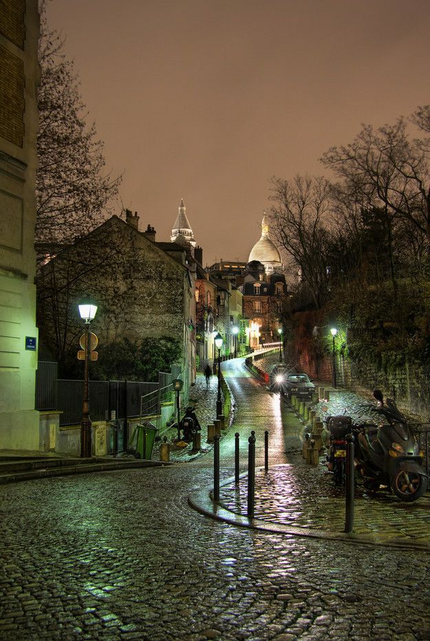 a cobblestone street at night with cars parked on the side and buildings in the background