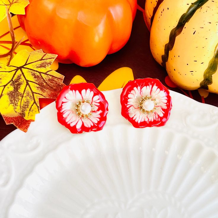 two red and white flower earrings sitting on top of a plate next to pumpkins