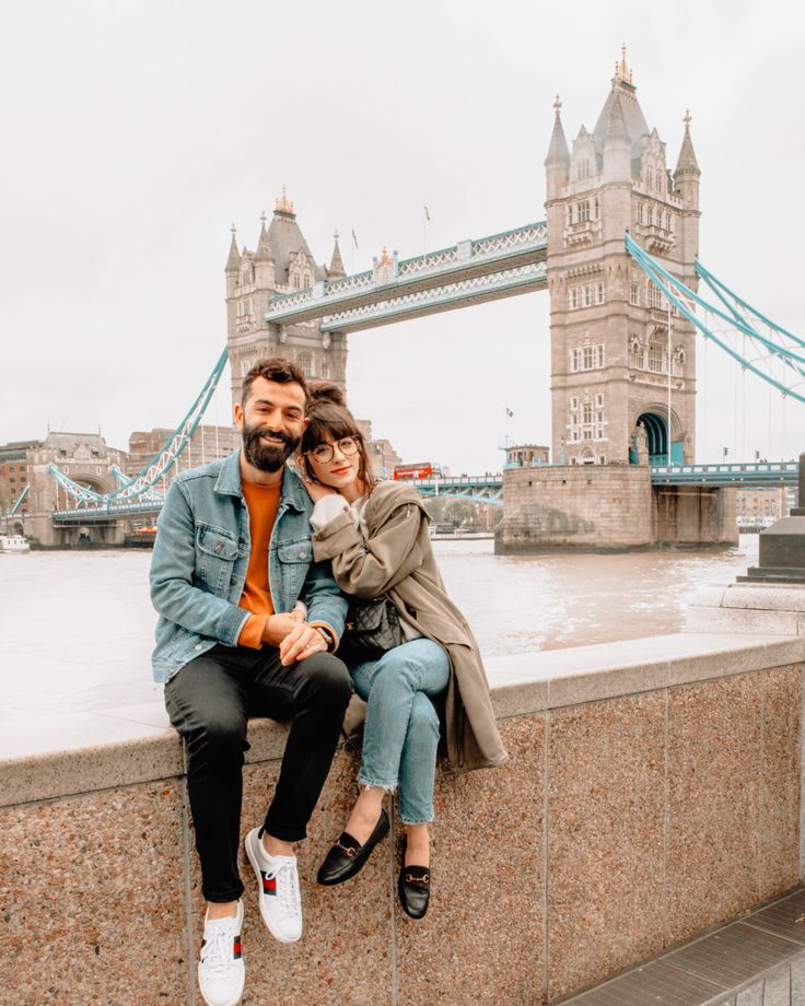 a man and woman sitting on a wall in front of the tower bridge