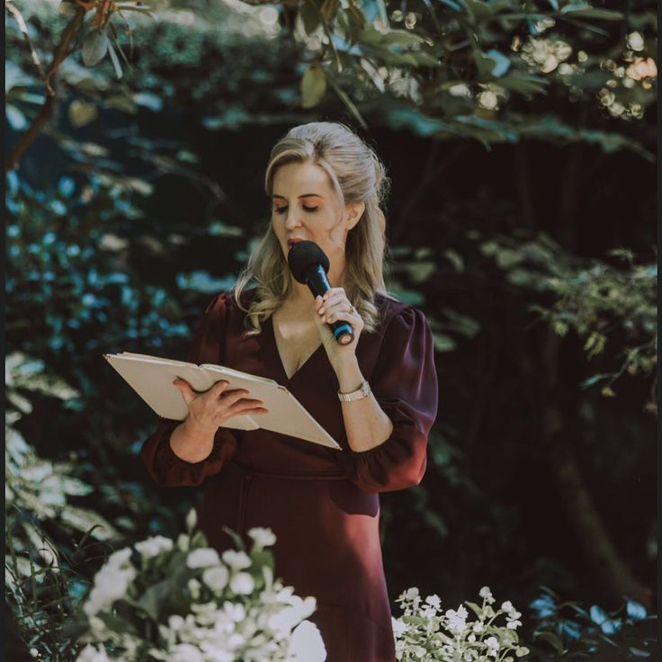 a woman in a red dress holding a microphone and reading a book while surrounded by flowers