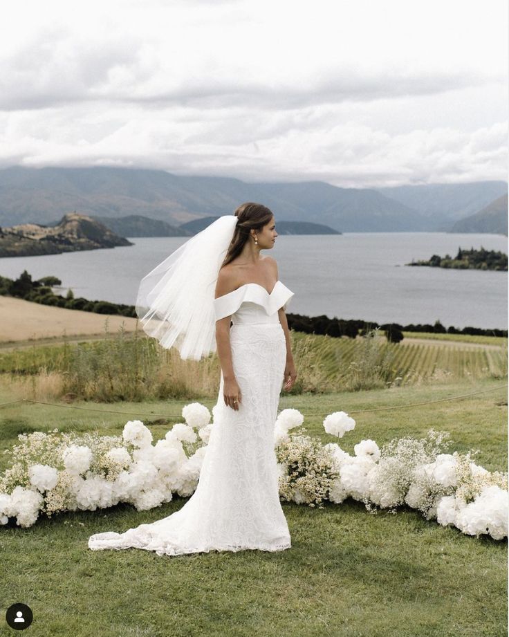 a woman in a wedding dress is standing near flowers and looking at the water from behind her