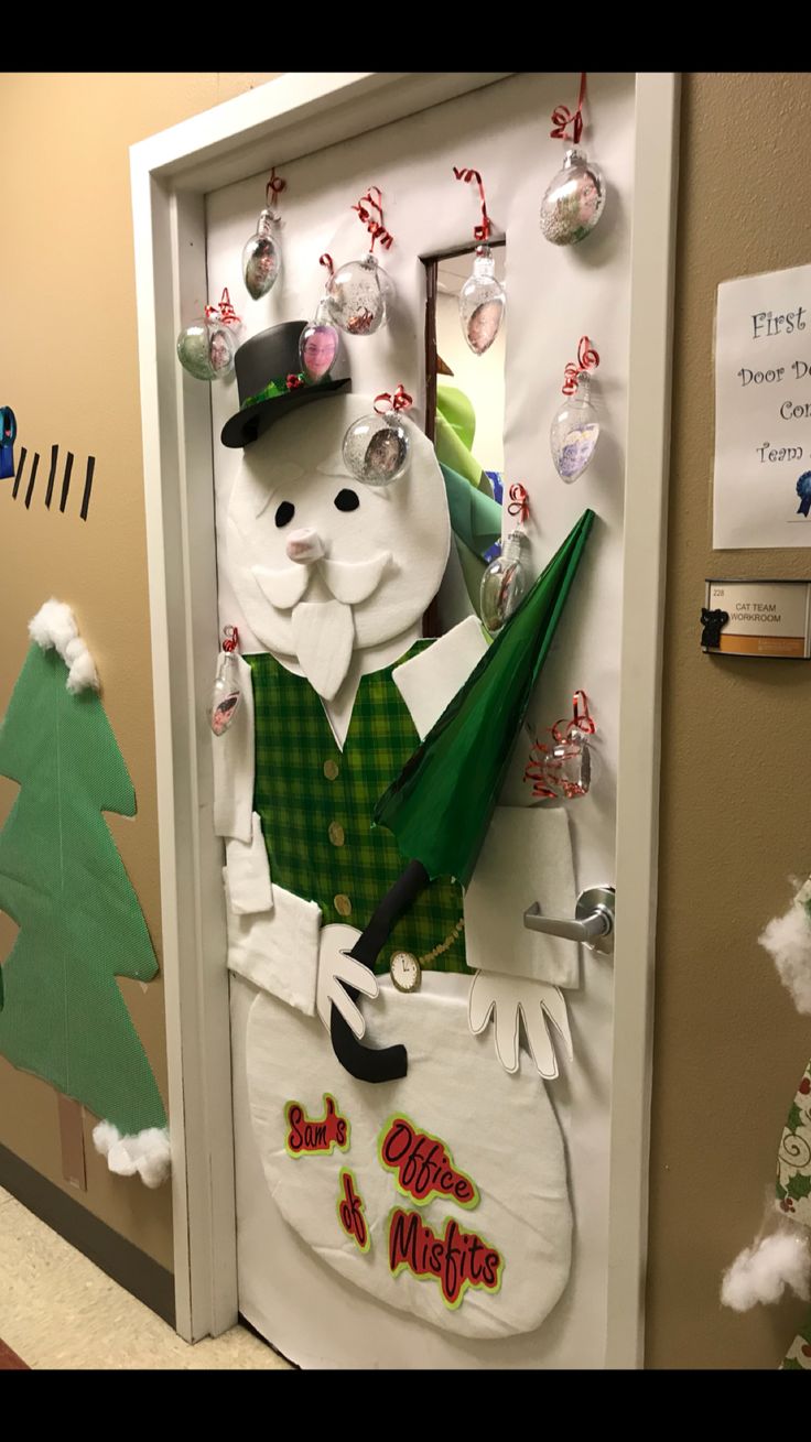 a door decorated to look like a snowman holding a green umbrella and hat, standing in front of a christmas tree