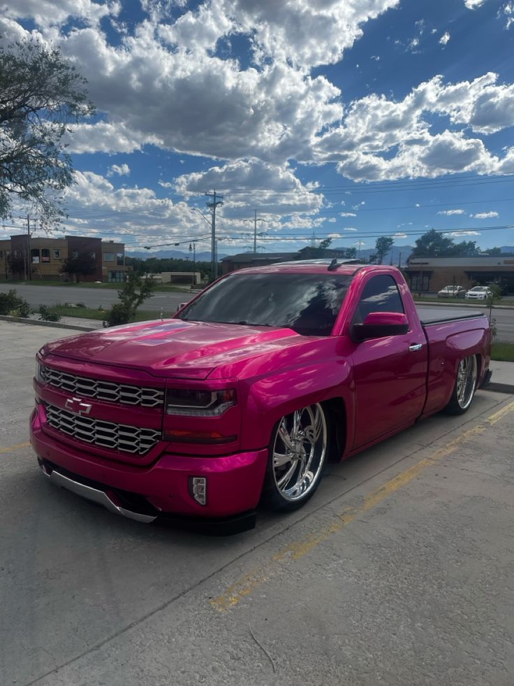 a pink truck parked in a parking lot next to a building with clouds above it