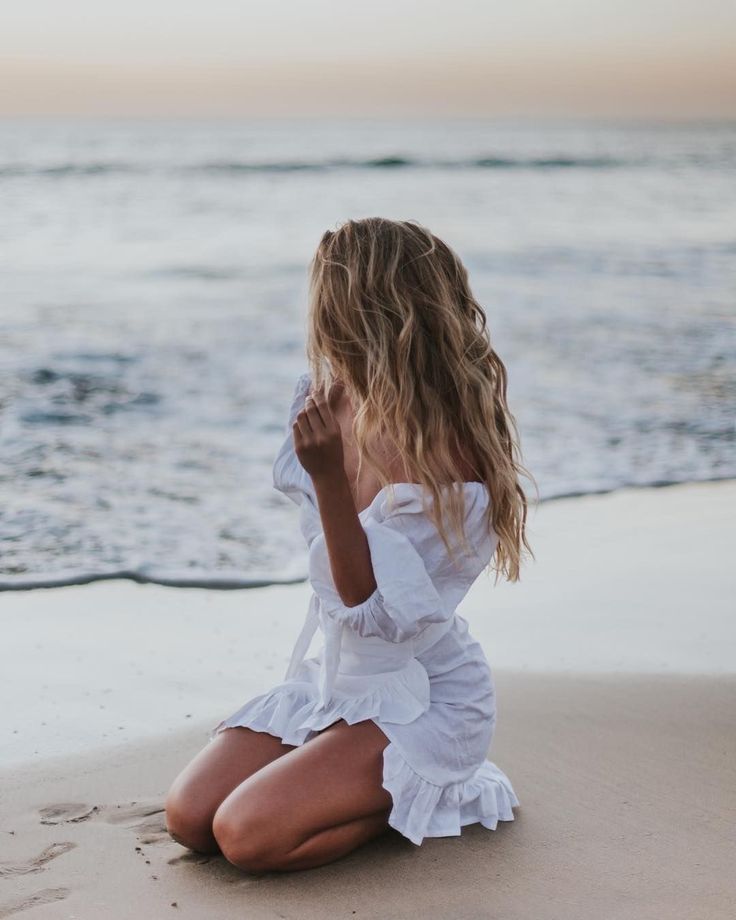 a woman sitting on top of a sandy beach next to the ocean with her hand in her mouth