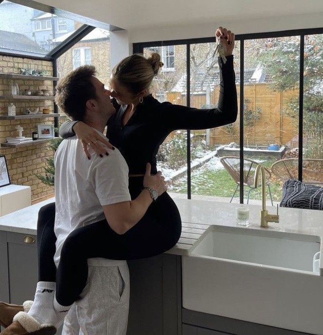 a man and woman standing in front of a kitchen window, one holding the other up