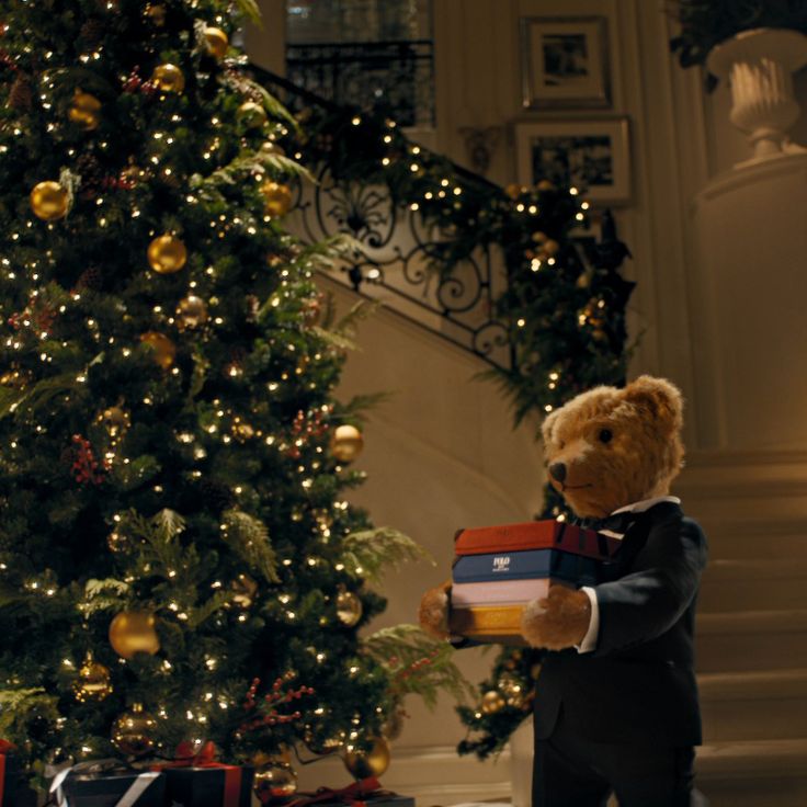 a teddy bear in a tuxedo is holding presents near a christmas tree