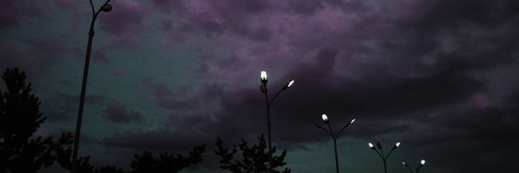 some street lights are lit up in the night sky with purple clouds behind them and trees