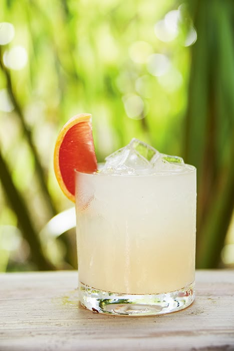 a drink with an orange slice on the rim sitting on a wooden table in front of some trees