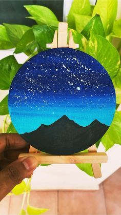 a hand holding up a small painting with mountains in the background and stars on it