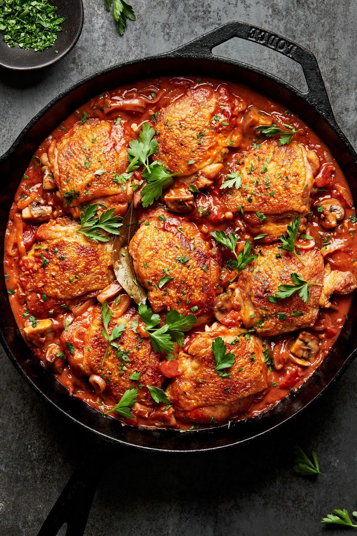 a pan filled with chicken and tomato sauce on top of a gray table next to some parsley