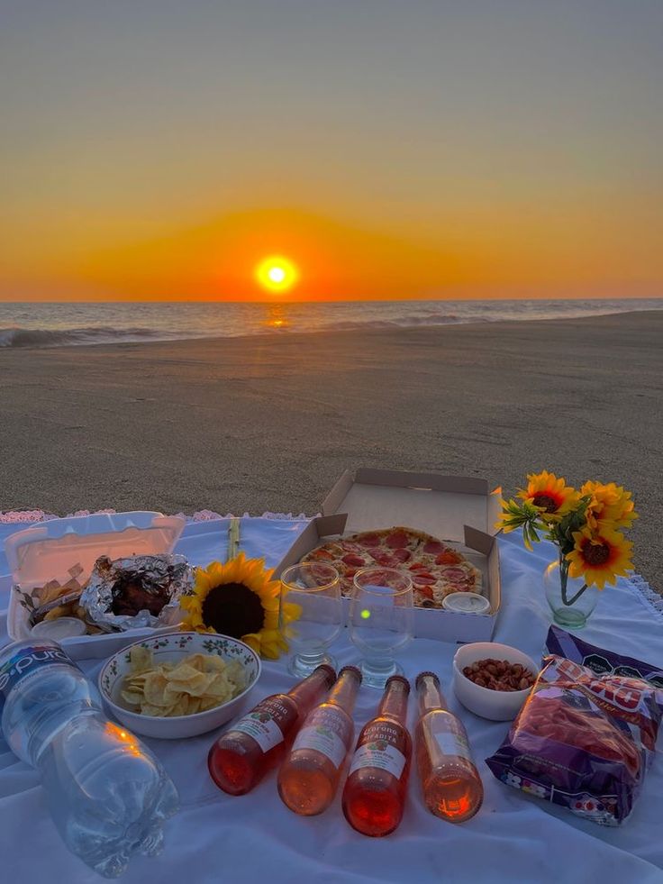 the sun is setting at the beach with food and drinks on the table next to it