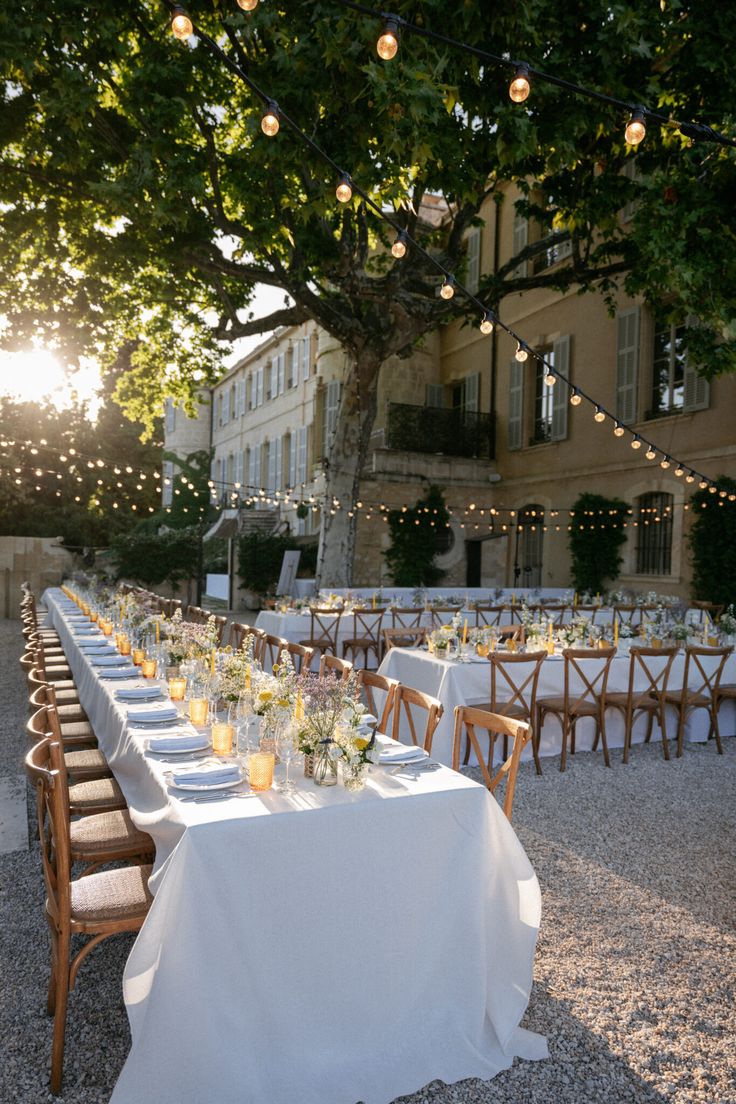 a long table set up with white linens and place settings for dinner under the shade of a tree