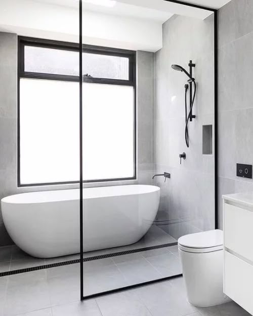 a white bath tub sitting next to a toilet under a window in a gray tiled bathroom