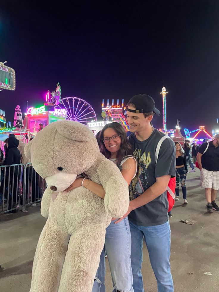 a man and woman standing next to each other holding a large teddy bear at an amusement park