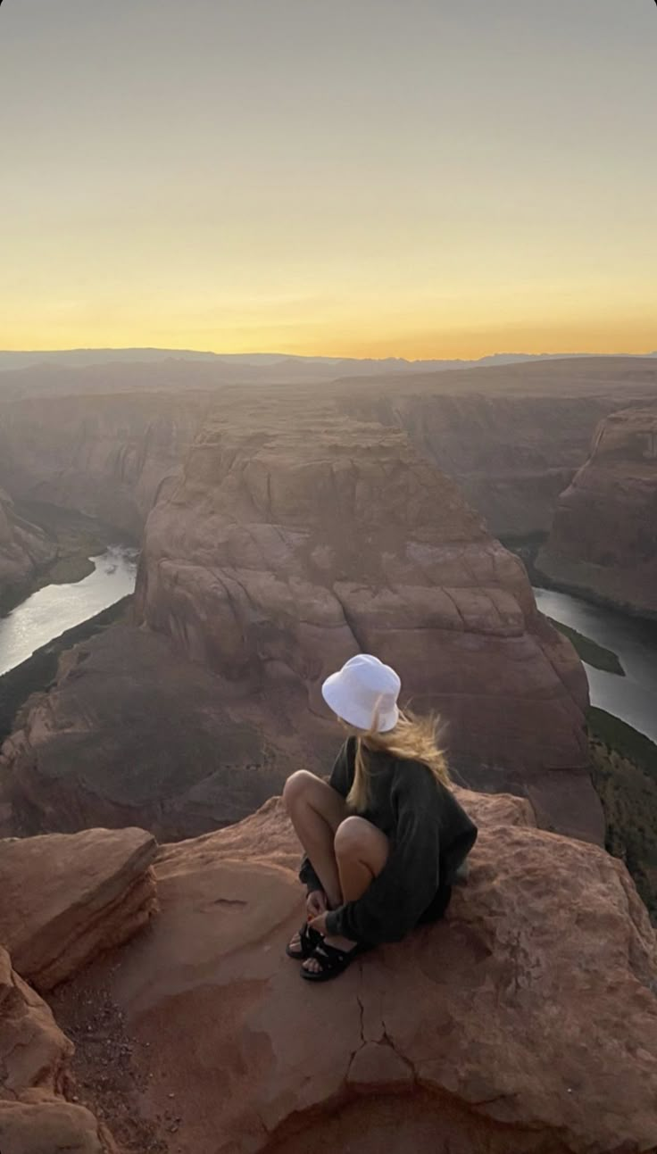 a woman sitting on top of a cliff next to a river and mountains at sunset