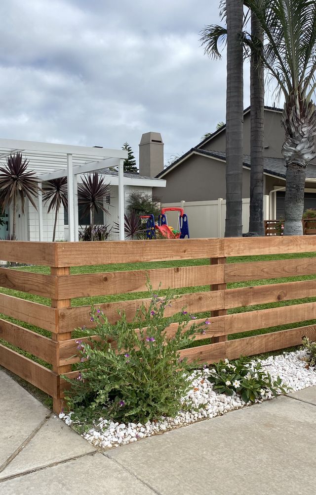 a wooden fence in front of a house with palm trees and flowers on the side