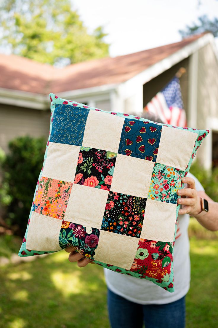 a person holding up a patchwork pillow in front of a house